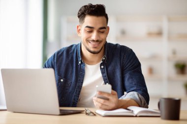 Neşeli Arap adam evde çalışma masasında otururken akıllı telefon kullanıyor, dizüstü bilgisayarda çalışıyor, yer kopyalıyor. Elinde cep telefonu olan mutlu, yakışıklı, orta doğulu bir adam.