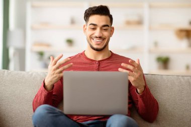 Olumlu Arap adam kız arkadaşıyla görüntülü konuşma yapıyor, evde dizüstü bilgisayar kullanıyor, Müslüman genç adam oturma odasında oturuyor, el kol hareketi yapıyor ve gülümsüyor.