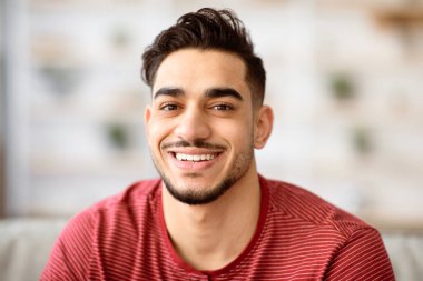 Evdeyken kameraya gülümseyen neşeli, yakışıklı bir Arap 'ın yakın plan portresi, kopyalama alanı. Pozitif genç, orta doğu sakallı adam evde poz veriyor, günlük kıyafetler giyiyor.