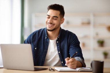 Memleketten dizüstü bilgisayarla çalışan neşeli bir Arap adam. Not defterine bakıyor ve notlar alıyor. Boşluğu kopyalıyor. Ofisteki çalışma masasında oturan mutlu orta doğulu genç adam.