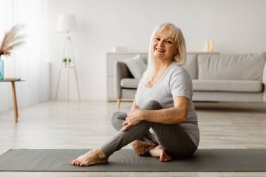 Ev Fizyoterapi Konsepti. Oturma odasında gülümseyen, kıdemli bayan egzersizi yapan, yoga minderinde oturan, poz veren ve kameranın keyfini çıkaran egzersiz yapan kadın portresi. Boş Kopyalama Alanı, Bulanık Arkaplan
