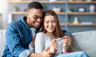 Pozitif gebelik testi yapan ve kanepede kucaklaşan mutlu çok kültürlü çift, romantik siyah erkek ve beyaz kadın ebeveyn olmaktan memnunlar, kanepede otururken sarılıyorlar, yakınlaşıyorlar.