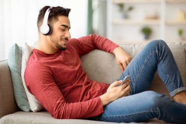 Rahat Arap adam evdeki koltukta dinlenirken cep telefonundan video izliyor, kablosuz kulaklık kullanıyor, kopyalama alanı kullanıyor. Neşeli orta doğulu adam akıllı telefondan film izliyor.