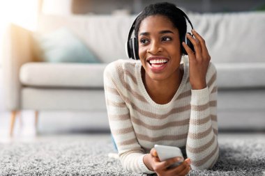 Telsiz kulaklıklı neşeli siyahi kadının yakın plan portresi, akıllı telefondan müzik dinleyen neşeli Afrikalı Amerikalı kadın oturma odasında dinlenirken favori şarkıların tadını çıkarıyor.