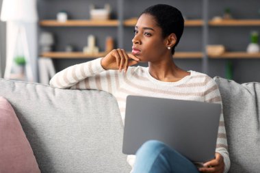 Evdeki koltukta dizüstü bilgisayarla oturan entellektüel Afrikalı Amerikalı kadının portresi, oturma odasında bilgisayarla çalışırken yeni kitabı düşünen düşünceli siyah kadın yazar,