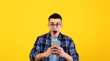 Gözlüklü yakışıklı genç adam turuncu stüdyo arka planında şok içinde akıllı telefonuna bakıyor. Cep telefonlu şaşırtıcı bir milenyum adamı tanıtım satışına ya da büyük bir indirime hayran kaldı.
