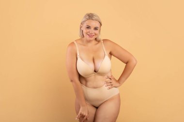 Artı Boyut Kadın. Güzel kıvrımlı kadın iç çamaşırlı pozu bej rengi stüdyo arka planında duran, kameraya gülümseyen, uzay kopyası olan çekici, iri yarı kadın portresi.