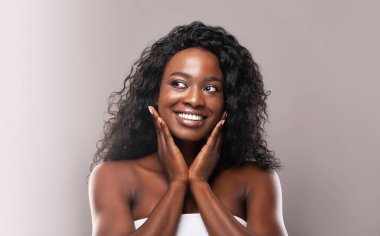 Gençleştirme konsepti. Güzel Afrikalı Amerikalı kadın mükemmel tenine yanaklarından dokunuyor ve gri arka plandan kameraya bakıyor.