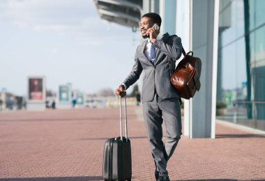 Modern bir havaalanının dışında bavullu profesyonel bir siyah adam arama yapıyor. Ruh hali dinamik ve odaklanmış, çağdaş bir ortamda iş seyahatinin özünü yakalıyor..