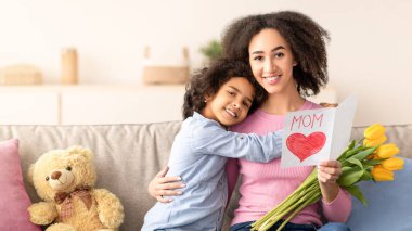 Bağlanma ve şefkat. Pozitif siyah kız portresi anneyi kartla selamlıyor ve bir demet taze sarı laleyle, gülümseyen kadını kucaklıyor. Mutlu bir aile kanepede oturup anneler gününü kutluyor.