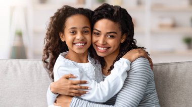 Güzel, genç, siyah bir anne ve tatlı bir kız evde oturuyor, oturma odasında kanepede oturuyor ve sarılıyor, yakından portre çekiyor. Annelik, ebeveynlik, anne-kız kavramı.