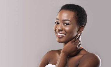 Güzellik ve Kişisel Bakım Konsepti. Güzel bir Afrikalı kadın boynuna dokunuyor, gülümsüyor ve kameraya gri arka plandan bakıyor, panorama
