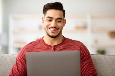 Laptop kullanan gülümseyen orta doğulu bir gencin yakın plan fotoğrafı, kopyalama alanı. Evinden çalışan neşeli Arap adam, oturma odasında koltukta oturuyor, modern not defteri kullanıyor. Uzak görev konsepti