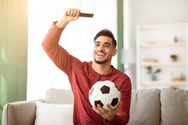 Evde TV 'de futbol maçı izleyen, elinde uzaktan kumandayla gezen, mutlu bir Arap futbol fanatiği oturma odasında oturmuş televizyon seyrediyor, maçtan zevk alıyor.