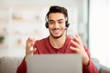 Neşeli Arap adamın evden görüntülü konuşma yapması, kulaklık ve dizüstü bilgisayar kullanması, defter ekranına bakması, gülümsemesi ve el kol hareketi yapması. Telekomünikasyon ve modern teknoloji kavramı