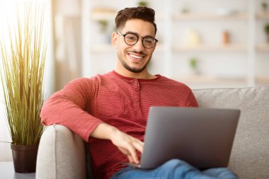 Evde çalışan, not defteri kullanan, oturma odasındaki koltukta oturan kameraya neşeyle gülümseyen, serbest çalışan, gözlüklü genç bir Arap. Bilgisayarı olan bir ortadoğulu.