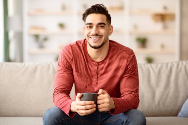 Rahat bir Arap adam kanepede oturur, çay ya da kahve içer, evin içinde. Orta Doğulu genç adam elinde fincanla evde vakit geçiriyor ve kameraya gülümsüyor.