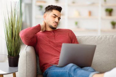 Evde dizüstü bilgisayar kullanırken boyun spazmı geçiren genç bir Arap adam oturma odasında kanepede oturuyor ve kaslarını ovuyor. Ortadoğulu adamın boynuna rahatsız edici bir durum yüzünden kramp girdi.