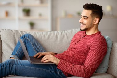 Orta Doğulu serbest çalışan, modern dizüstü bilgisayarla koltukta oturan ve klavyede yazan serbest çalışan bir adamın yan görüntüsü, kopyalama alanı. İnternette sörf yapan ya da arkadaşlarıyla sohbet eden mutlu bir Arap.