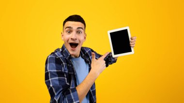 Heyecanlı hipster çocuk tableti işaret ediyor turuncu arka planda boş ekran, uygulama alanı ya da web sitesi tasarımı. Pozitif milenyum adamı reklamın için dokunmatik bloknotu gösteriyor.