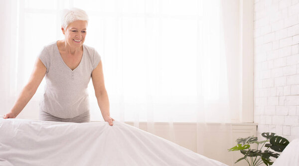 Пожилая женщина делает кровать и организует комнату утром, панорама, копировальное пространство
