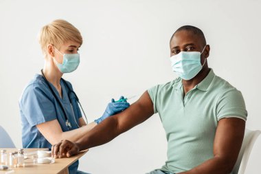 Hastalıklar, tıbbi bakım, bilim ve aşı kampanyası. Koruyucu maskeli ve eldivenli yetişkin kadın doktor masadaki Afro-Amerikan erkeğe ampul, ilaç ve şırınga enjekte ediyor.
