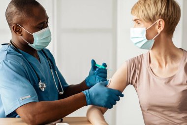 Salgın hastalık, covid-19 aşısı. Hasta, Afro-Amerikan üniformalı yetişkin erkeğe grip ya da COVID iğnesi yapan profesyonel bir doktor ya da hemşire koruyucu maskeli olgun bir kadını aşılıyor.
