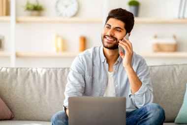 Serbest yaşam tarzı. Neşeli Arap adam cep telefonuyla konuşuyor laptopla çalışıyor, evdeki rahat kanepede oturuyor. Adam iş sorularını tartışıyor. Uzak mobil iletişim ve uzak iş kavramı