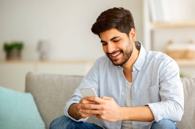 Neşeli Arap adam akıllı telefonuyla evdeki rahat koltukta oturuyor, arkadaşlarıyla mesajlaşıyor, cep telefonuyla dinleniyor ve yeni uygulamaları kontrol ediyor, mobil iletişimden keyif alıyor ya da sosyal ağları tarıyor.