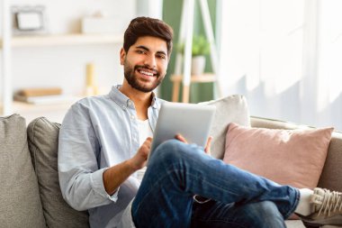 Boş zaman kavramı. Arap adam dijital tablet kullanıyor, internette geziniyor ve oturma odasındaki rahat kanepede dinlenirken sosyal ağları kontrol ediyor. Erkek alışverişi ya da online çalışma