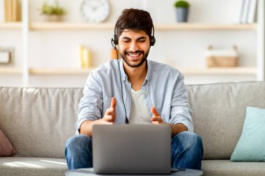 Video konferansı. Kulaklık takmış Arap bir adam dizüstü bilgisayarında, evden uzakta çalışıyor, kanepede oturuyor ve kameraya el kol hareketi yapıyor. Çevrimiçi iletişimi ve uzaktan çalışmayı seven adam