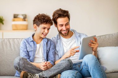 Mutlu baba ve oğul tablet bilgisayar kullanıyor, video izliyor ya da online oyunlar oynuyor, evde birlikte vakit geçiriyor, rahat bir koltukta oturuyor. Boş aktiviteler, modern teknoloji konsepti
