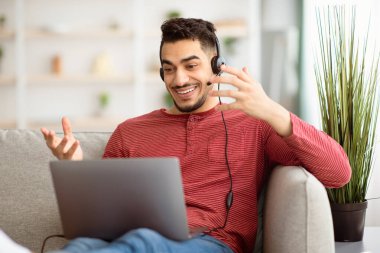 Evinden çalışan neşeli bir ortadoğulu, dizüstü bilgisayarla koltukta oturmuş, kulaklık takıyor. Arap adam müşterileri veya meslektaşlarıyla görüntülü görüşme yapıyor, el kol hareketi yapıyor ve konuşuyor, boşluğu kopyalıyor