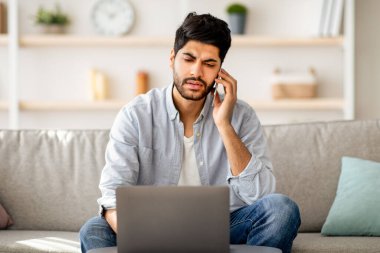 Uzaktan kumanda. Endişeli Arap serbest çalışanı cep telefonuyla konuşuyor, dizüstü bilgisayar kullanıyor, evdeki kanepede oturuyor. Uzaktan çalışan ve kurumsal telefon görüşmesi yapan bir adam.