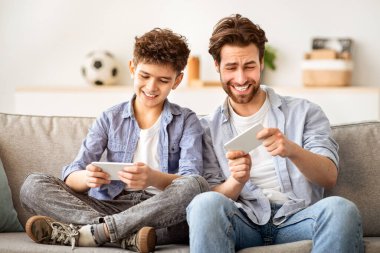 Eğlence ve rahatlık. Baba oğluyla online video oyunlarında akıllı telefonlarda yarışmak oturma odasında kanepede yan yana oturmak. Aile birlikte vakit geçiriyor. İki nesil, ortak hobi