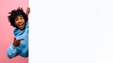 Heyecanlı Afrikalı Amerikalı genç boş bir posterin arkasına saklanıyor ve pembe stüdyo arka planındaki reklam tasarımın için bir maket hazırlıyor. Coşkulu siyahi genç adam boş pankart sunuyor, panorama