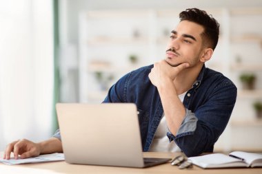 Bin yıllık düşünceli Arap adam ofiste oturuyor ve kopyalama alanına bakıyor, dizüstü bilgisayarla çalışıyor, yaratıcı fikirler arıyor. Çevrimiçi iş arayan yakışıklı bir ortadoğulu. Kariyer hayali kuruyor.