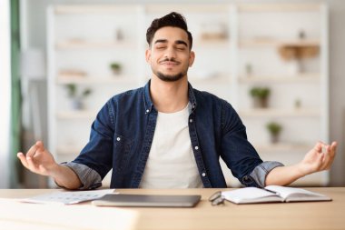 İş kavramında stres atmak. Arap adam çalışma masasında oturuyor, bilgisayarı kapalı, gözleri kapalı, Zen jesti ile el ele tutuşuyor, ofiste meditasyon yapıyor. Stresli bir günün ardından erkek çalışanın sakinleşmesi