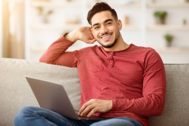 Mutlu Arap adamın yeni dizüstü bilgisayarının keyfini çıkarırken, oturma odasında kanepede oturmuş kameraya gülümserken. Memleketten çalışan neşeli bir orta doğulu adam. Defter kullanıyor, kopyalama alanı kullanıyor.