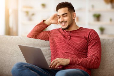 Mutlu Arap adam kanepede oturup dizüstü bilgisayar kullanıyor, çöpçatan sitesinde sohbet ediyor, bilgisayar ekranına bakıyor ve gülümsüyor. Sosyal medyada sörf yapan genç ortadoğulu adam, boşluğu kopyala