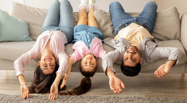 Upside down portrait of happy young family lying on sofa with heads down. Mother, father and daughter having fun, in modern living room interior. Go crazy together