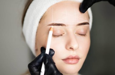 Profesyonel kozmetikçi, Güzellik Salonunda Kalıcı Tedaviden Önce Beyaz Kalemli Genç Kadının Kaşlarını Haritalamak, Büyülü Bölgeyi ve PMU işleminden Önce Alnını İşaretlemek,