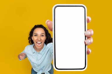 Harika bir teklif. Heyecanlı genç bayan elinde büyük bir cep telefonu ile boş beyaz model ekran aygıtını işaret ederek yeni mobil uygulamayı, sarı turuncu duvarı, yüksek açılı üst görüşü, kopyalamayı öneriyor.