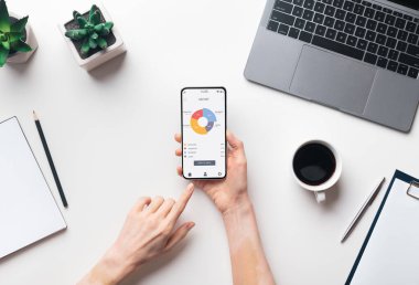 İş kadını elleri cep telefonunu elinde tutuyor. Çalışma masasında ticaret uygulaması, üst görünüm, yaratıcı imaj. Ofiste çalışırken akıllı telefondan finansal uygulama kullanan tanınmayan kadın yönetici