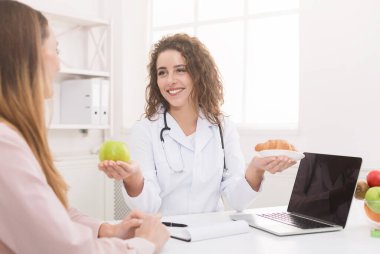 Kadın beslenme uzmanı hastaya doğru beslenmenin faydalarını anlatıyor, elma ve kruvasan tutuyor, abur cubur ve sağlıklı yiyecekleri karşılaştırıyor, kopyalama alanı yaratıyor.