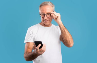 İnsanlar ve Teknoloji. Mutlu gülümseyen olgun bir adamın portresi akıllı telefon kullanarak ve dokunarak mavi stüdyo duvarının üzerinde gözlük takarak. Yetişkin erkek sohbet ediyor, sosyal medyada geziniyor.