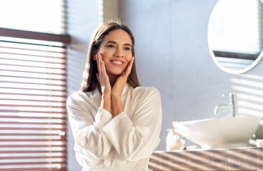 Güzellik Konsepti. Beyaz ipek bornoz giyen genç ve çekici bir kadın modern banyoda poz veriyor, güzel bir kadın yüzüne dokunuyor, evcil cilt bakımıyla eğleniyor.