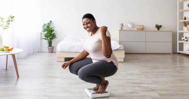 Heyecanlı kıvrımlı siyah kadın evde terazide oturuyor, EVET jesti yapıyor, zayıflatma diyeti ve panoramasından memnun. Aşırı neşeli ve iri yarı Afrikalı Amerikalı kadın kilo verme hedefine ulaşıyor.