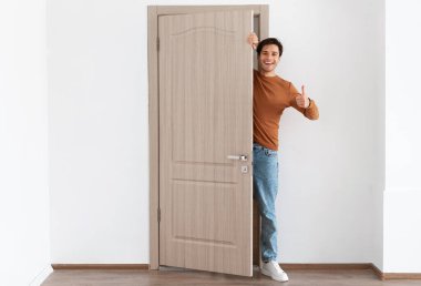 İyi teklif. Evinin kapısında duran neşeli genç adamın portresi, gülümseyen milenyum erkek ev sahibi aralık kapı tutuyor, dışarı bakıyor, ziyaretçiyi selamlıyor, işaret işareti gibi başparmaklarını gösteriyor.