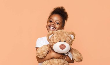 Şeftali rengi arka planda kahverengi bir ayıyı tutarken kıvırcık saçlı Afrikalı küçük kız gülümsüyor. O beyaz bir tişört giyiyor ve mutlu görünüyor..